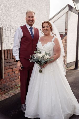 A newlywed couple stands together on their wedding day, with the bride in a white gown and groom in burgundy suit.