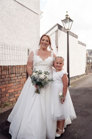 Bridesmaids wearing elegant white dresses carry bouquets while walking along a brick wall path.