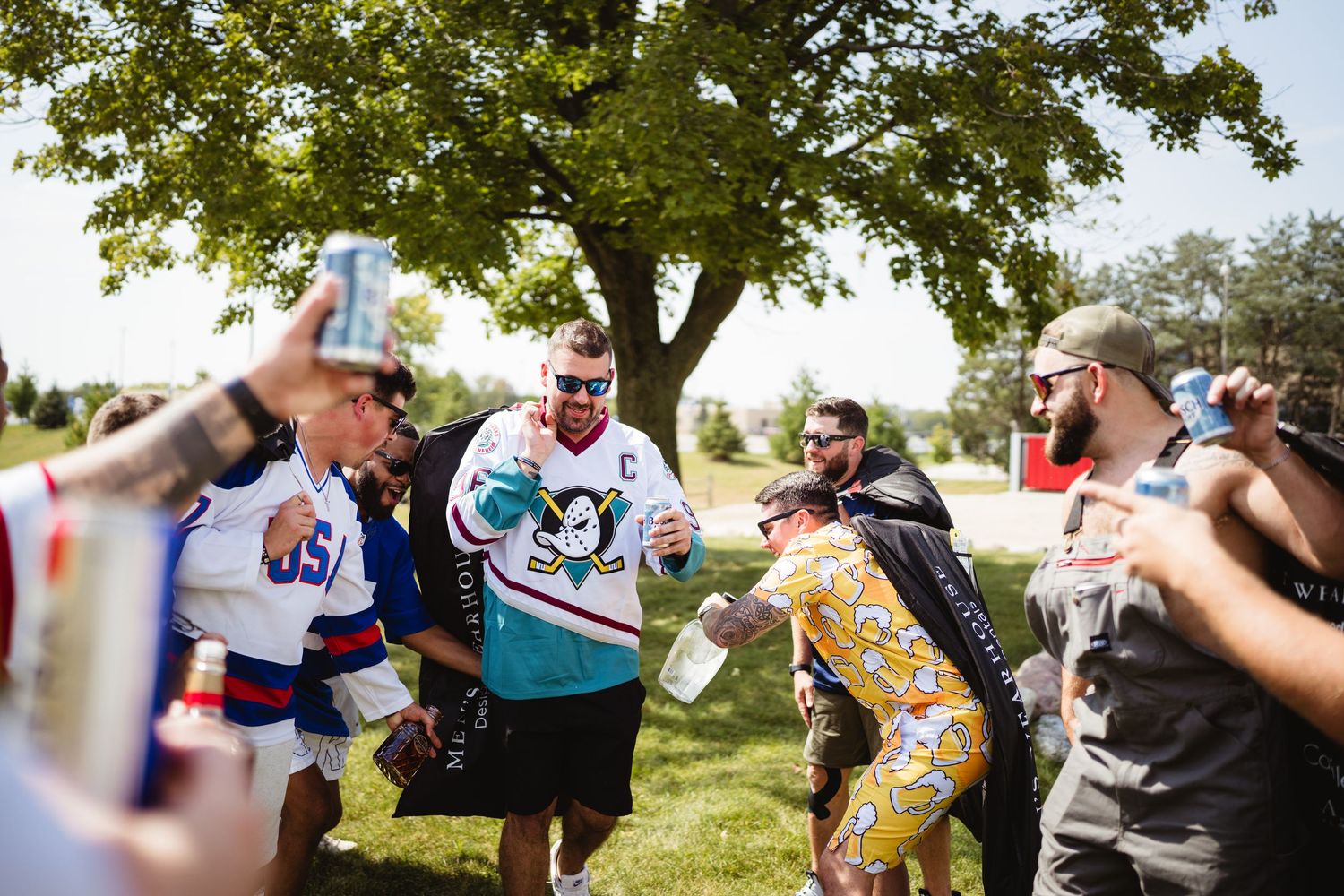 A group spraying celebratory champagne bottles outdoors under a large tree on a sunny day.