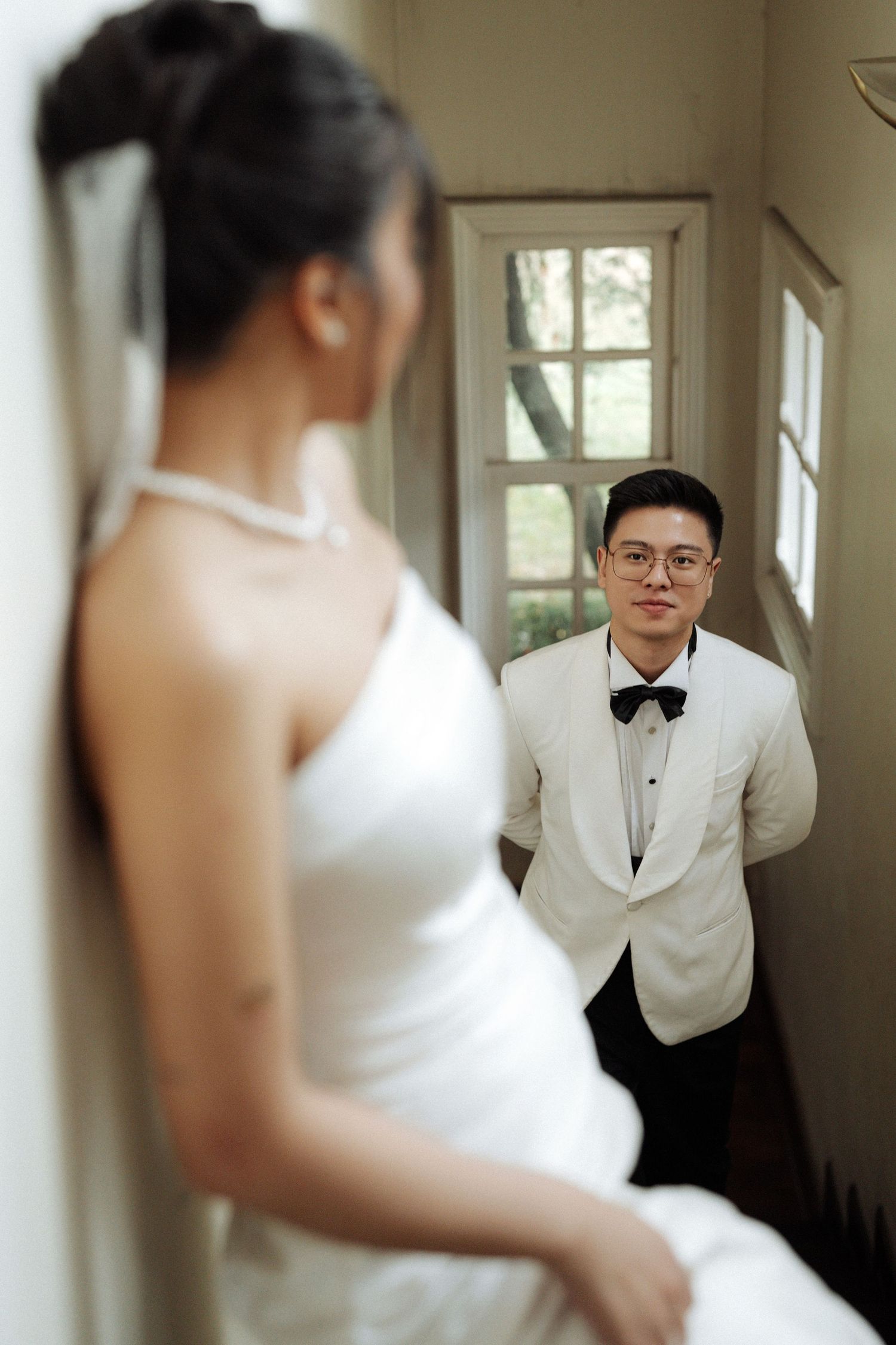 Groom gazes admiringly at bride leaning against a wall in a softly lit hallway portrait.