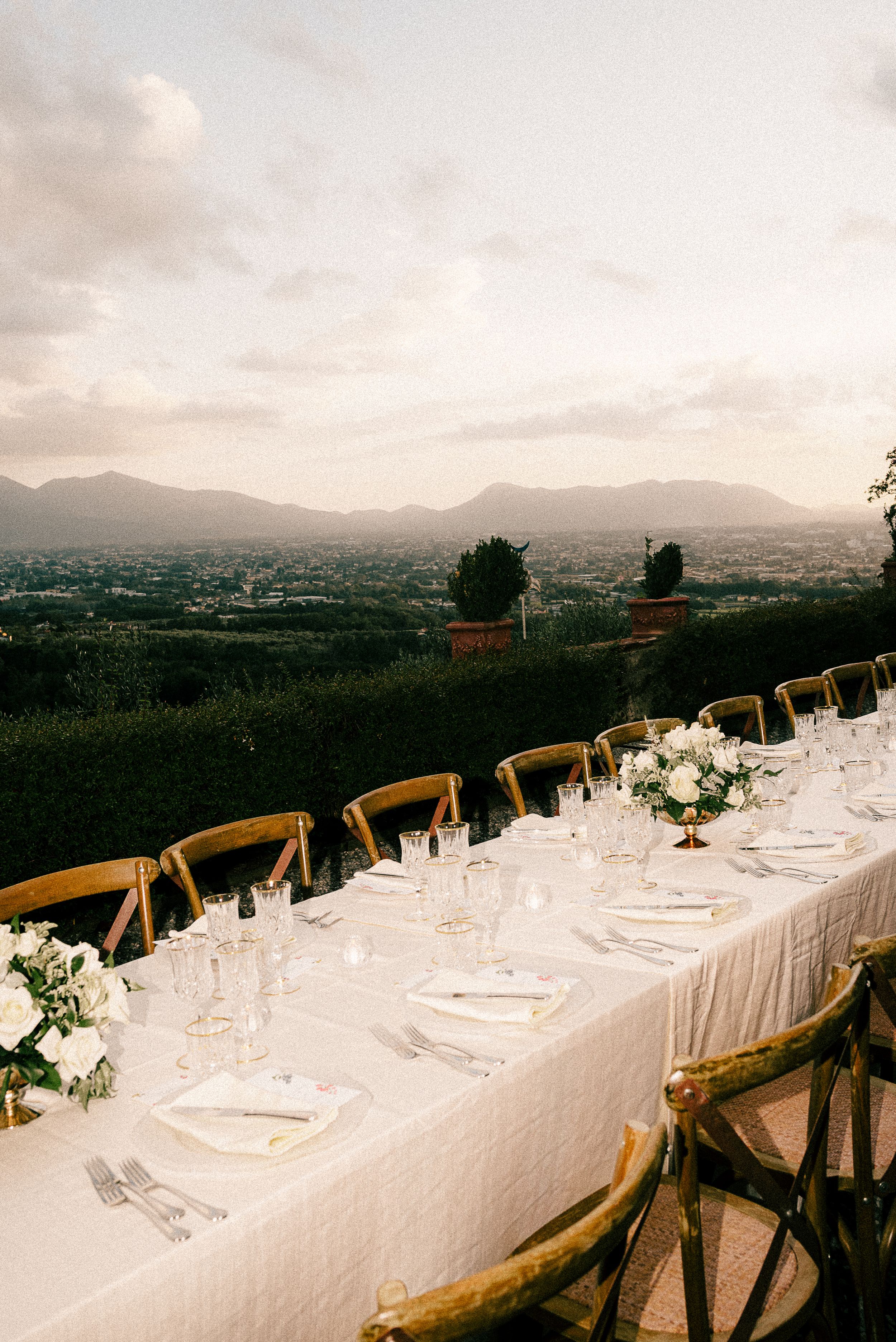 Elegant outdoor wedding reception tables set with white linens and wooden chairs against a scenic mountain backdrop.