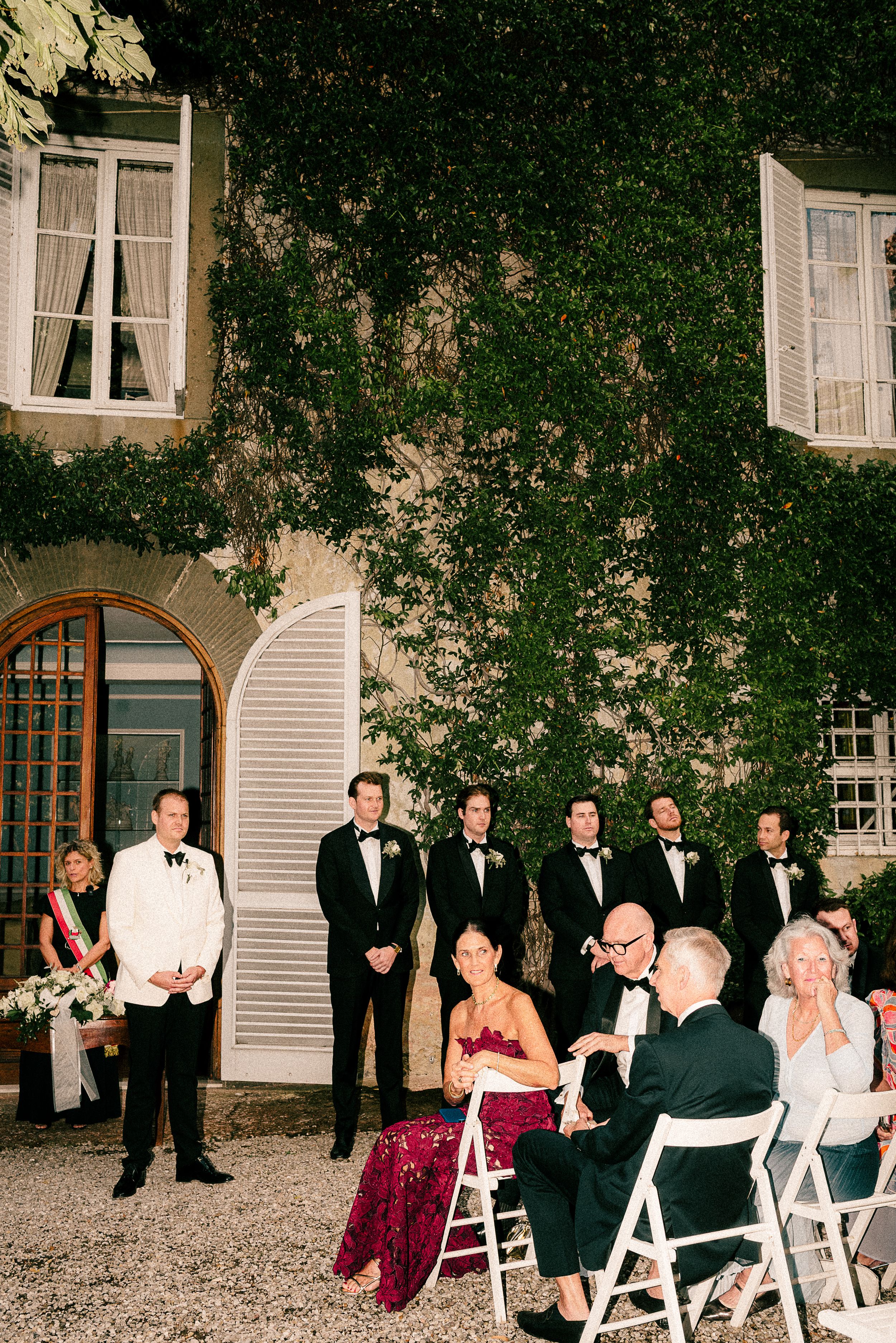 Formal wedding party members gather on a garden patio with elegant white chairs.