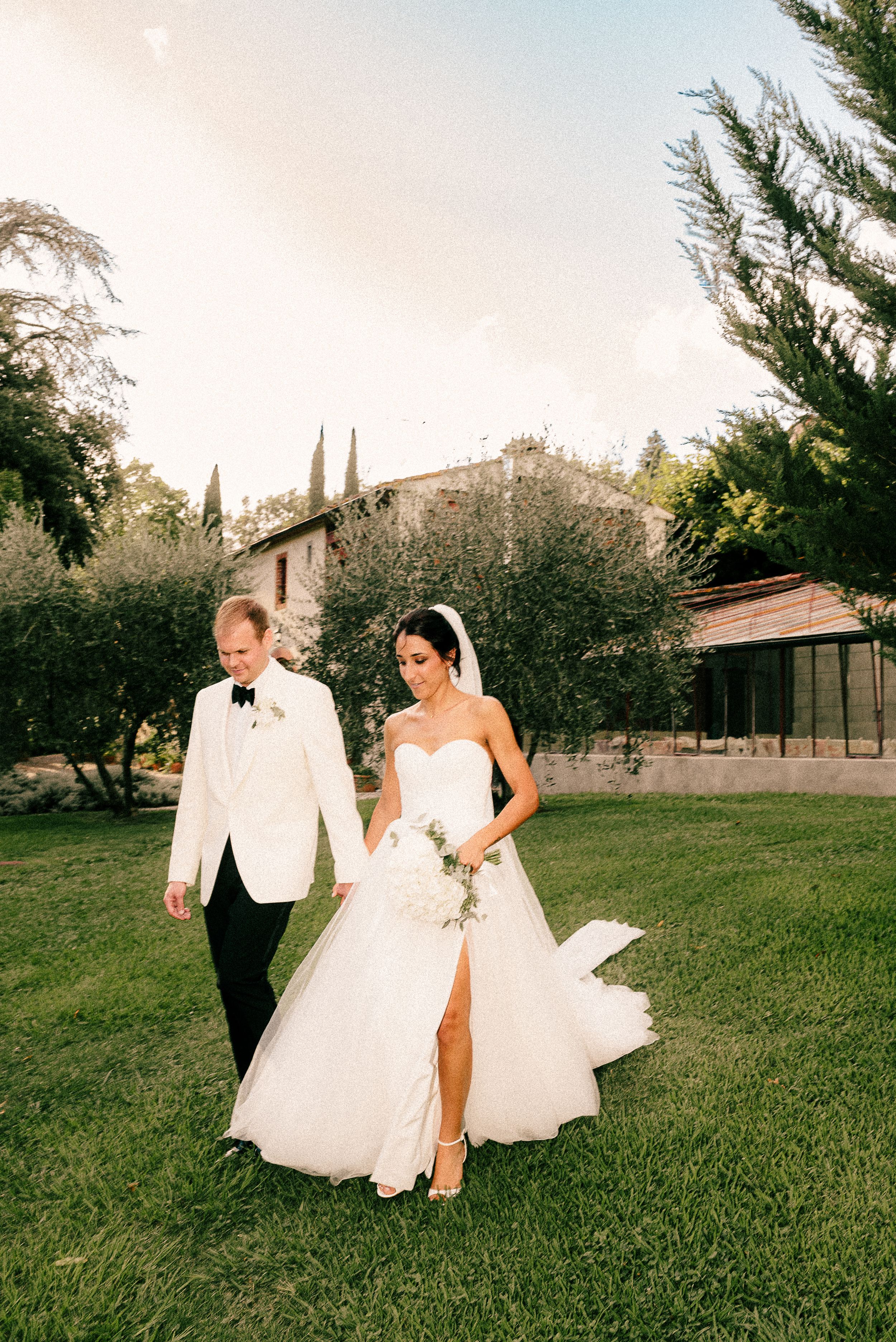 Couple strolls across a lush green lawn at sunset during their wedding celebration.