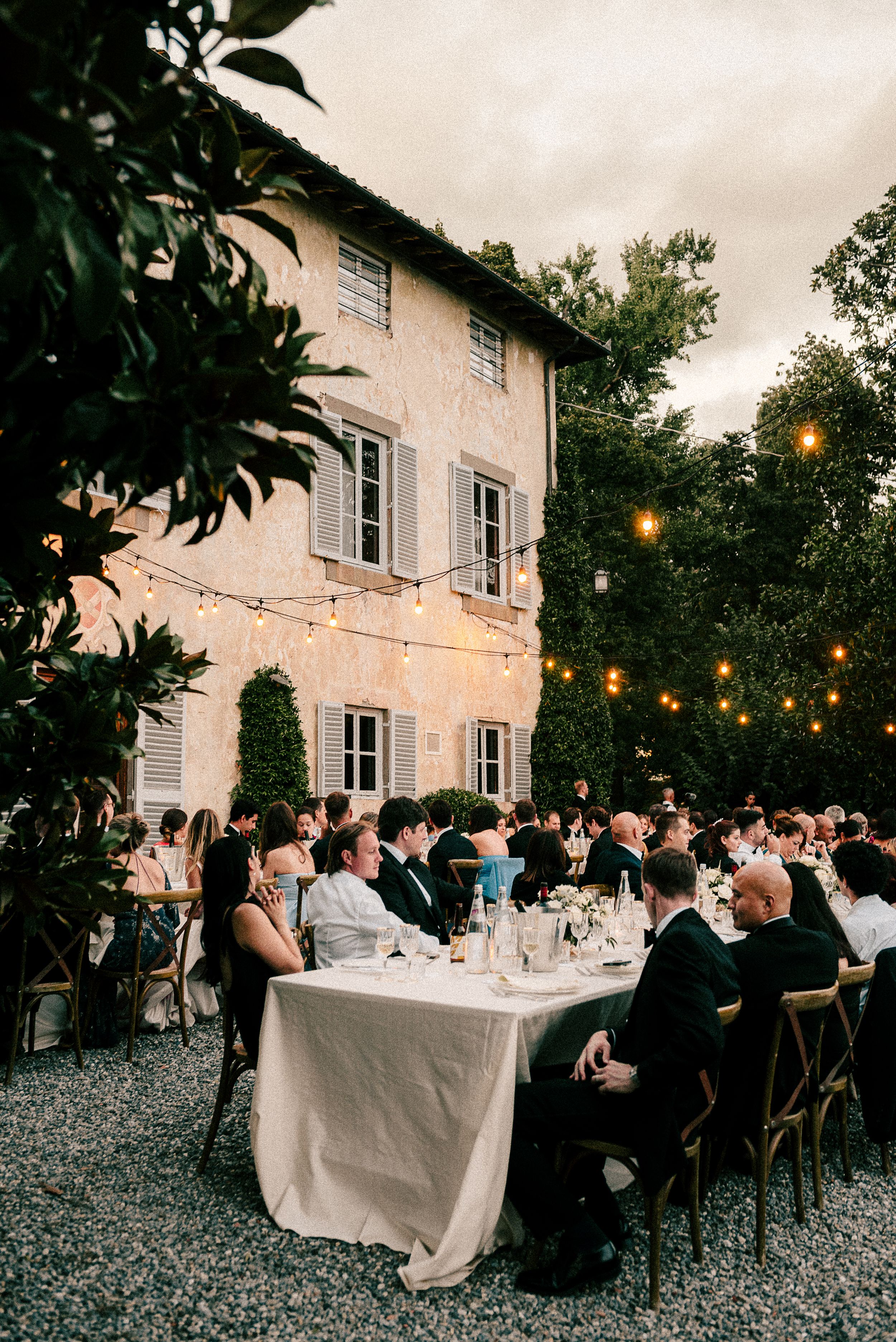 Outdoor wedding dinner setup with string lights and guests seated at long tables against a rustic villa backdrop.