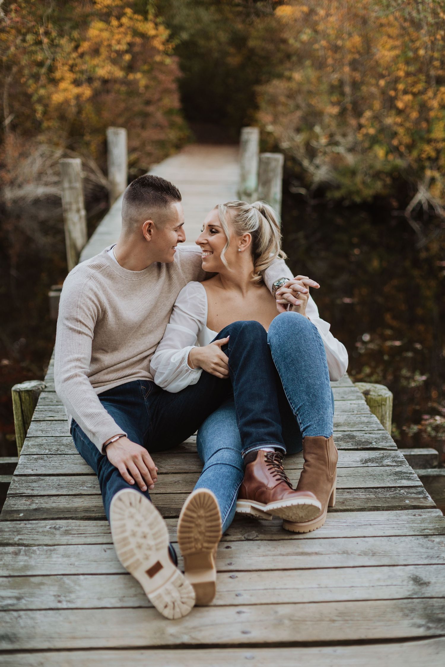 A couple sits together on a wooden dock surrounded by autumn foliage reflected in calm water below.