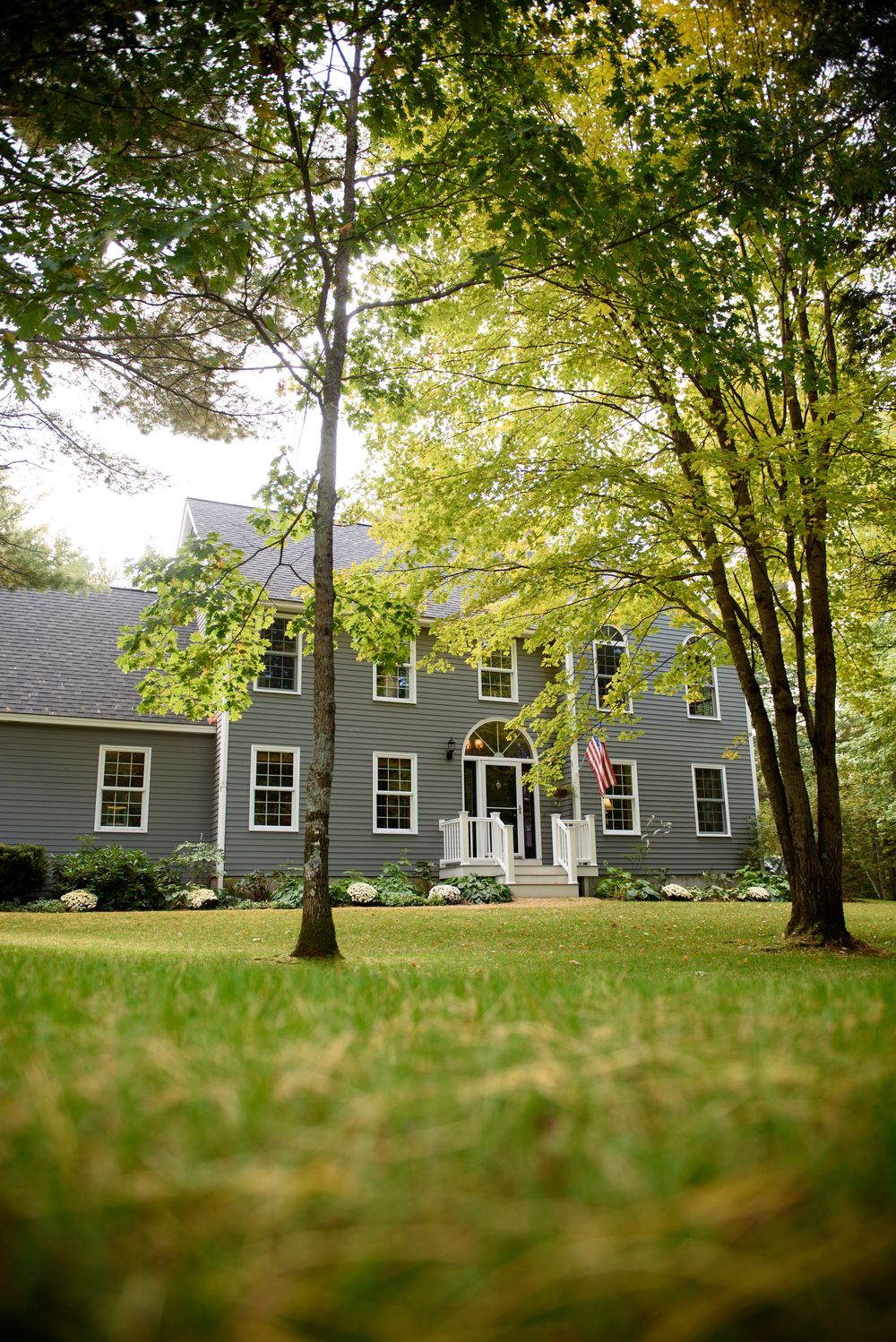 Gray colonial style house nestled among mature trees on green lawn.