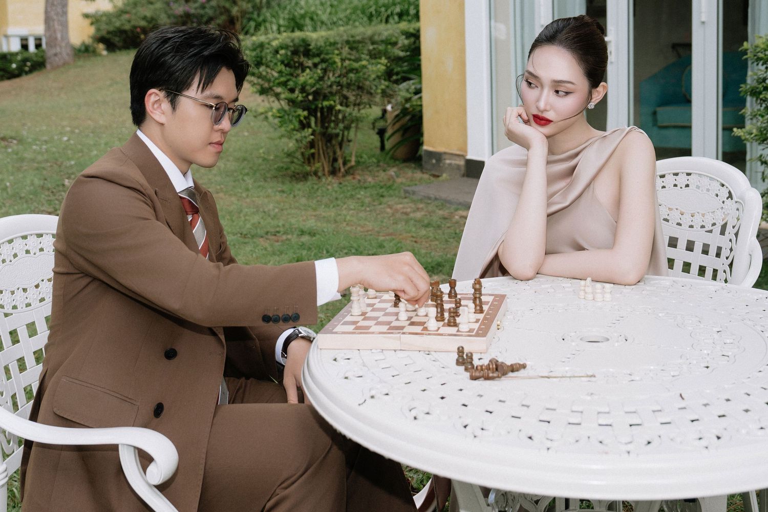 Elegant couple playing chess at a white outdoor table in a lush garden setting.