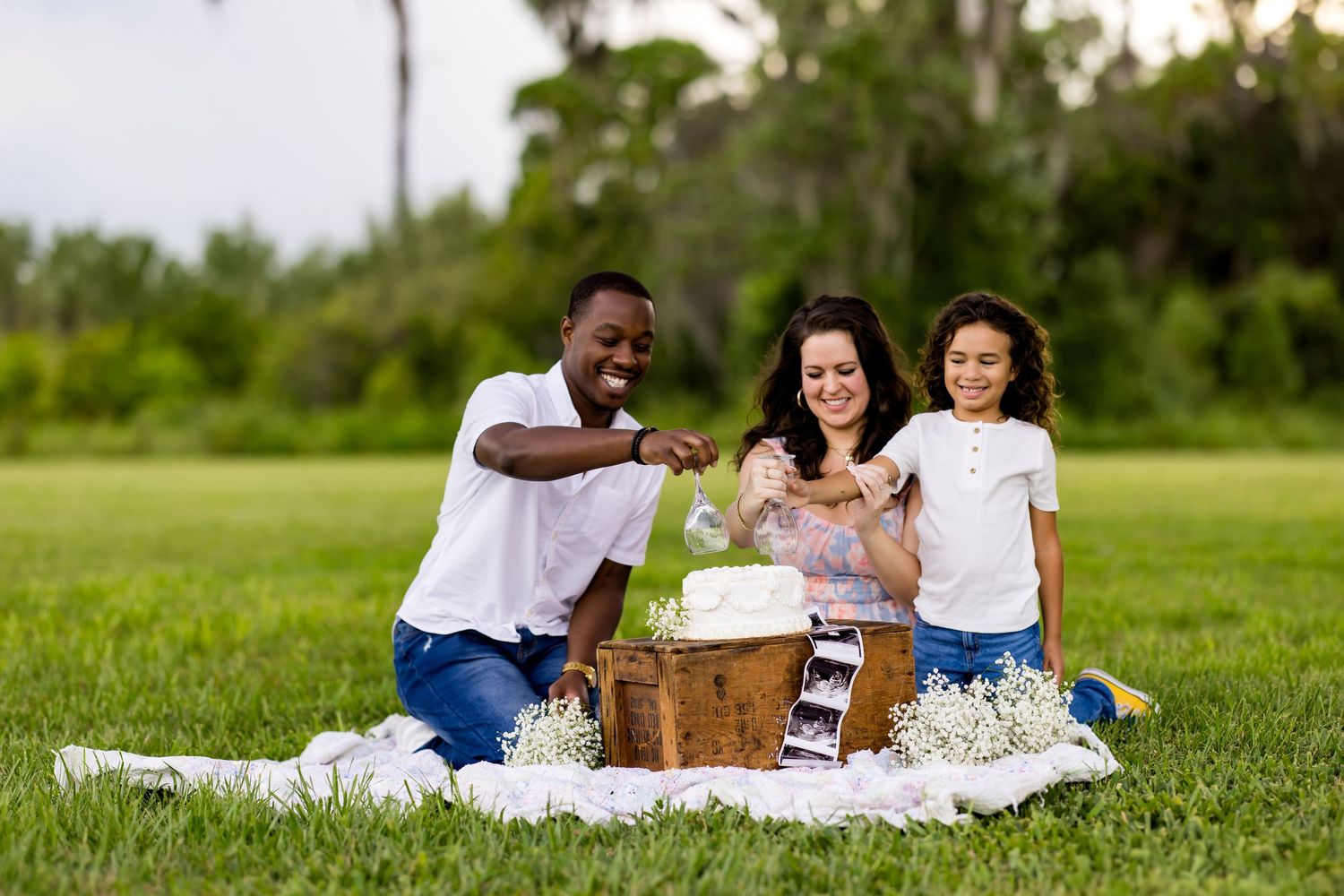 A family enjoys an outdoor picnic together on a white blanket in a lush green field during a sunny day.