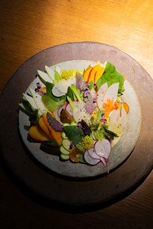 Artfully arranged fresh vegetables and edible flowers on a handcrafted ceramic plate under spotlighting.