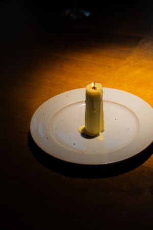 Single white candle centered on minimalist white plate under spotlight.
