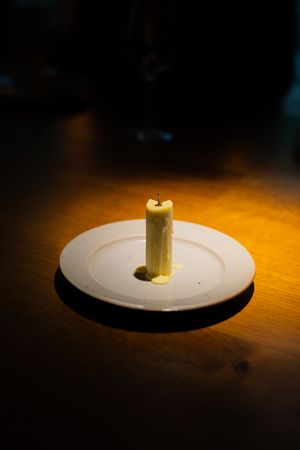 A single white candle burning on a plain white plate against a dark wooden surface.