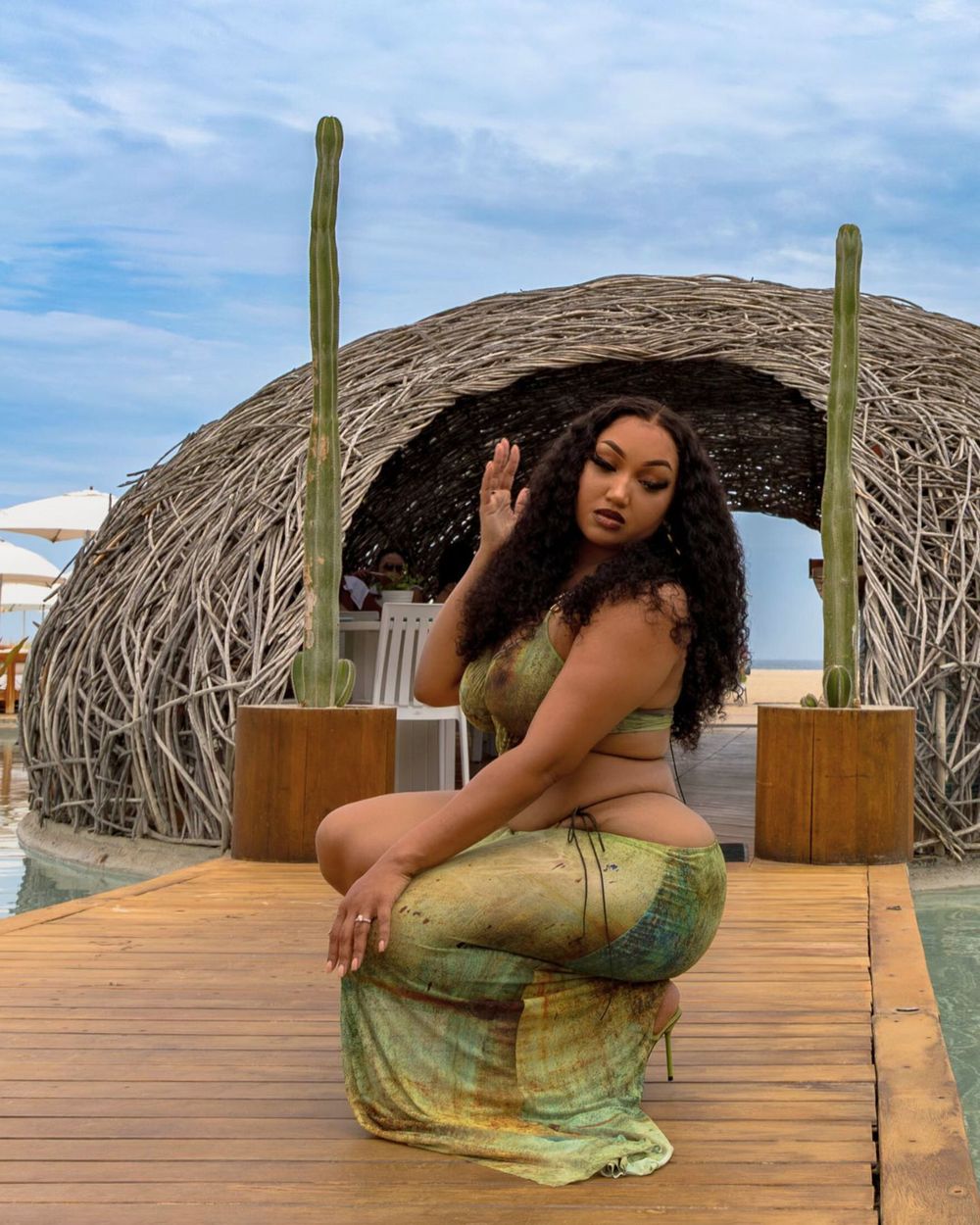 A figure in a flowing green dress poses gracefully on a wooden deck with desert cacti and domed structures in the background.