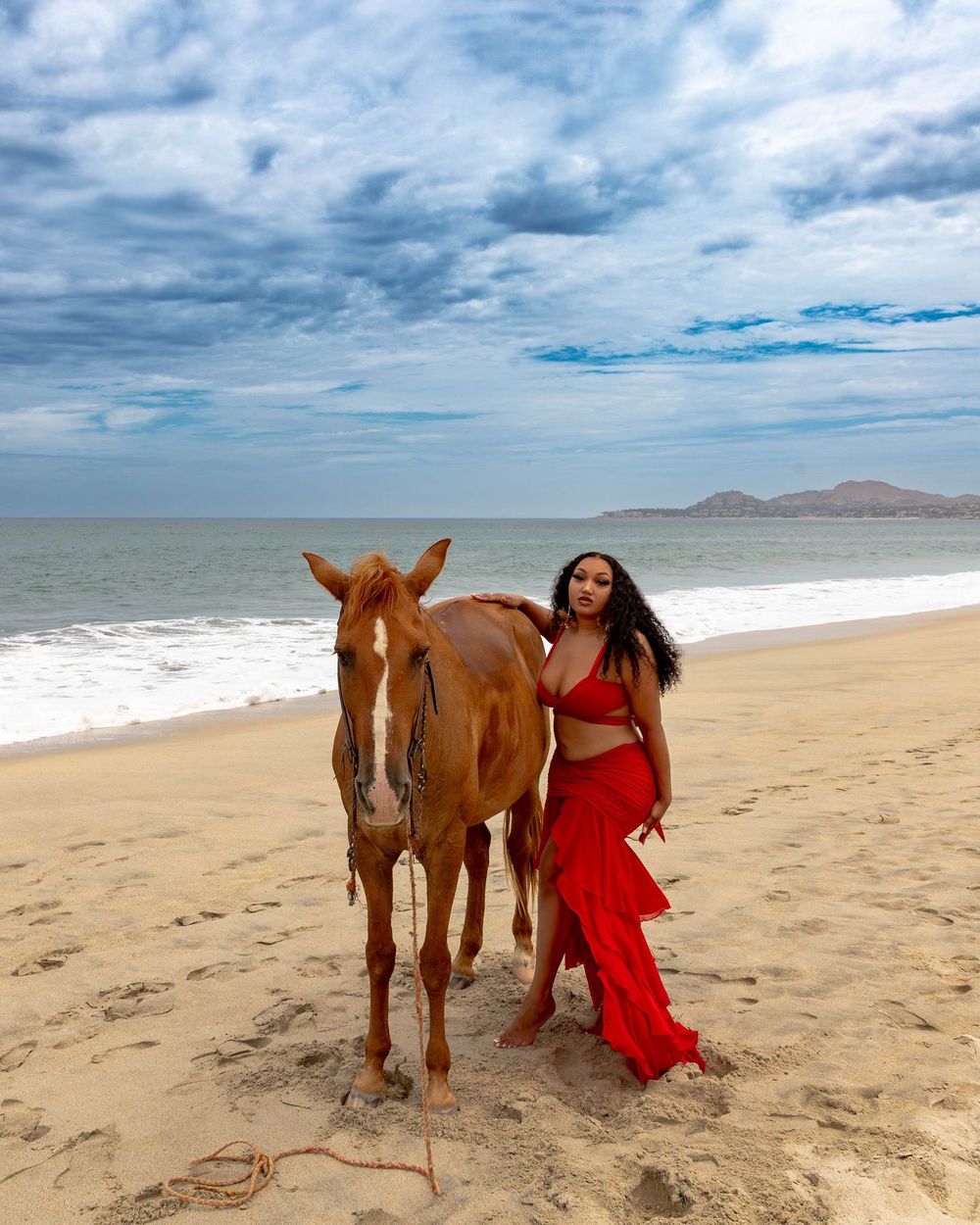 A scenic beach photoshoot featuring a horse and someone in a flowing red dress against a cloudy coastal backdrop.