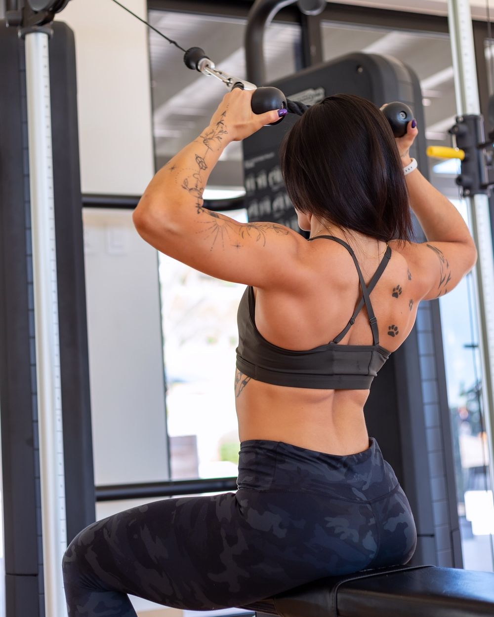 Person with dark hair and tattoos performs lat pulldown exercise at a gym using cable machine equipment.