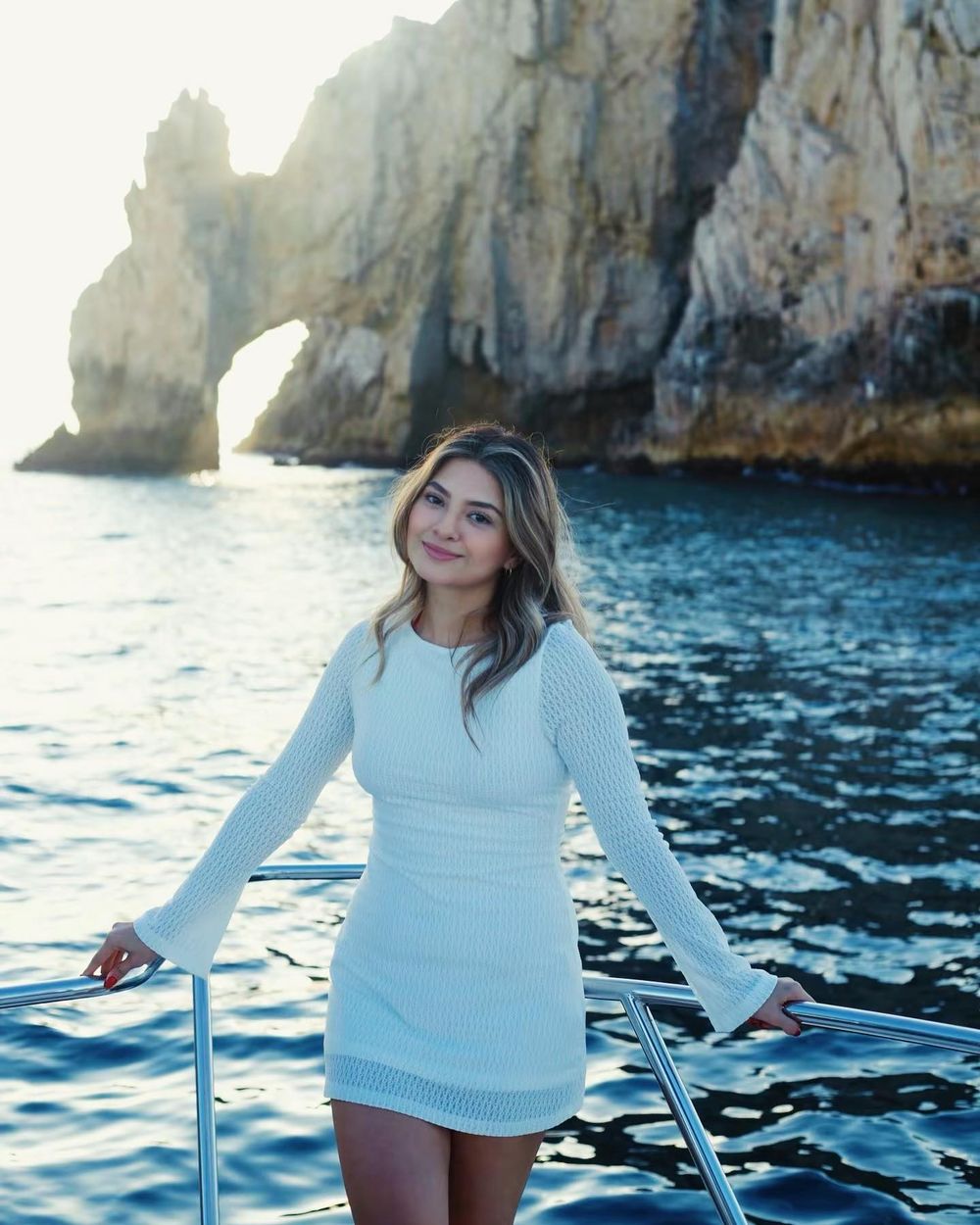 Woman in white sweater poses on a boat with rocky coastal cliffs and blue Mediterranean waters in the background.