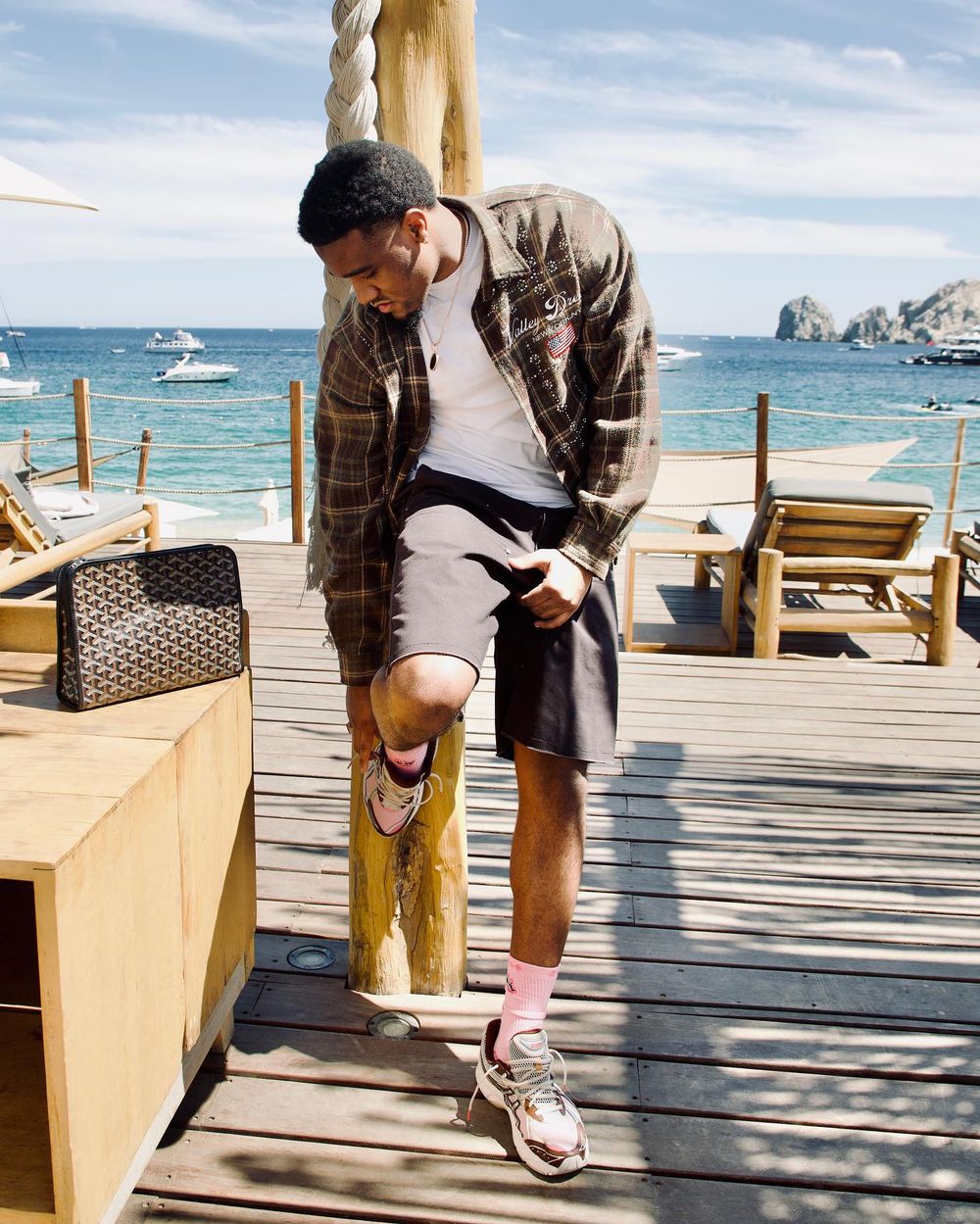 Person in plaid shirt and shorts kneeling on wooden boardwalk by ocean with boats and cliffs visible.