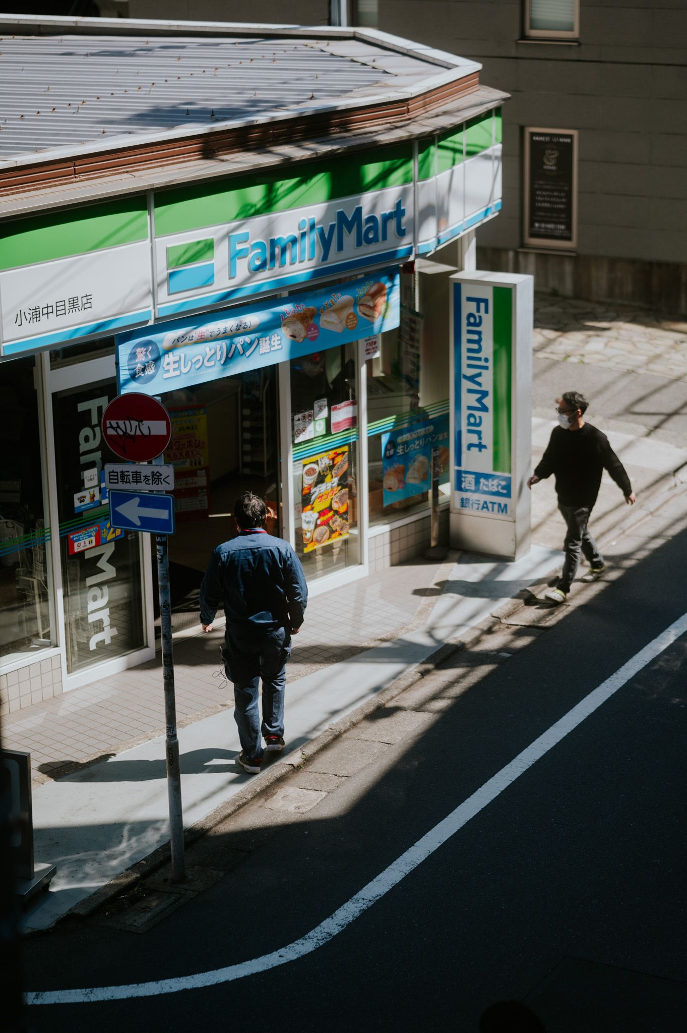 Shadowy figures walk past a convenience store storefront with blue and green signage in an urban setting during daytime.