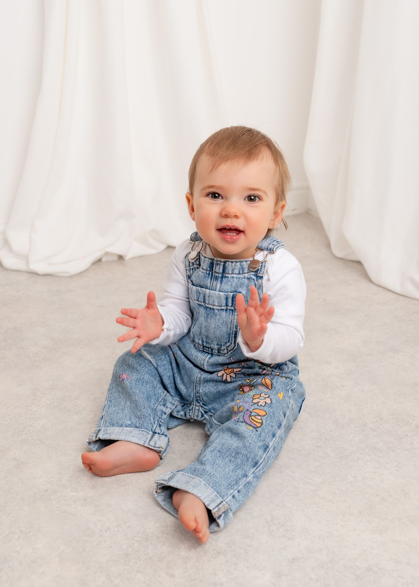 Baby in denim overalls sits and plays on white backdrop in progressive photo sequence at her 1st birthday photography session.