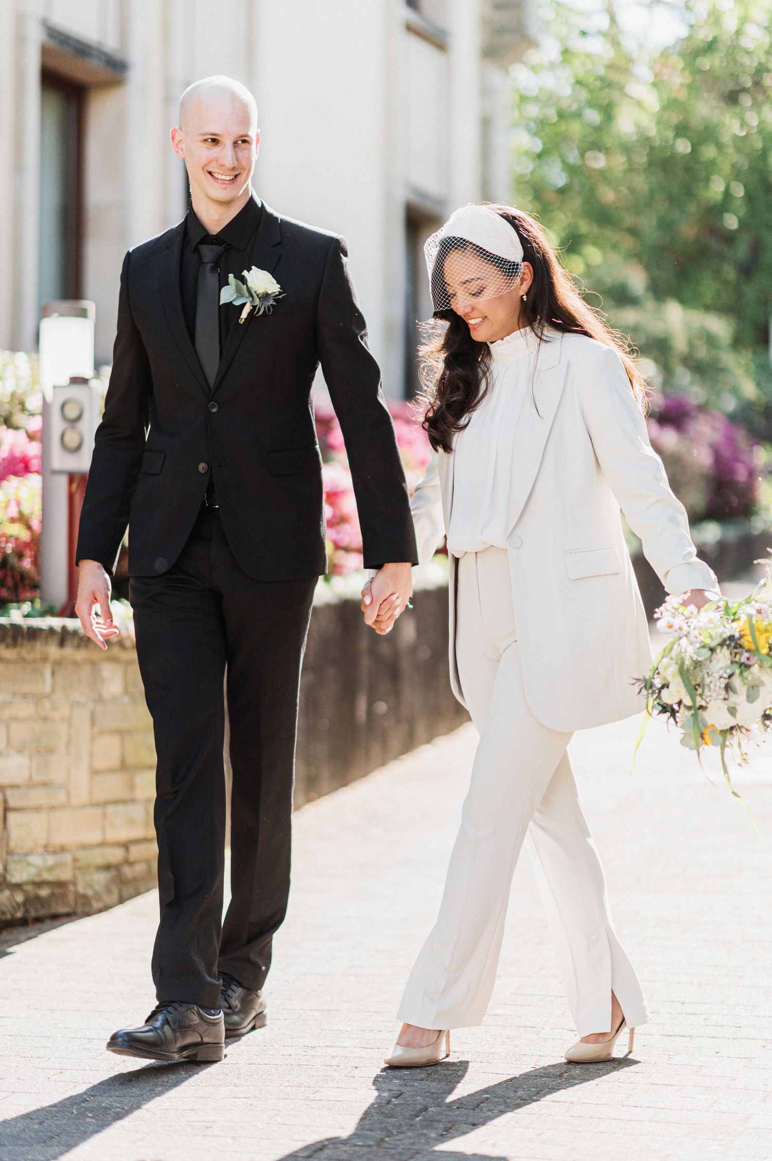 A couple walking together on a sunny day, wearing black and white formal outfits while holding hands.