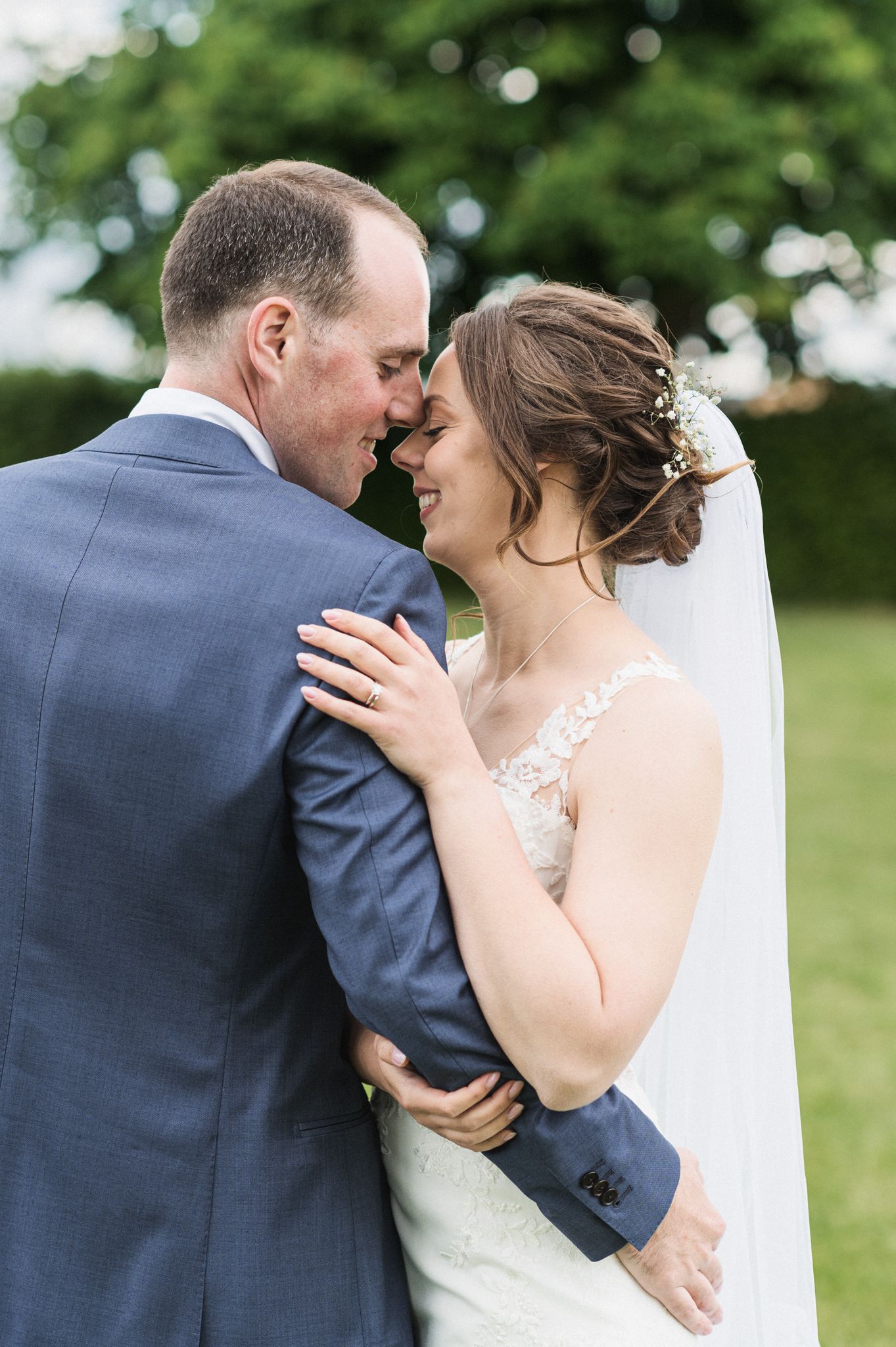 A tender moment between newlyweds outdoors as they embrace, with the bride in white lace and groom in navy suit.