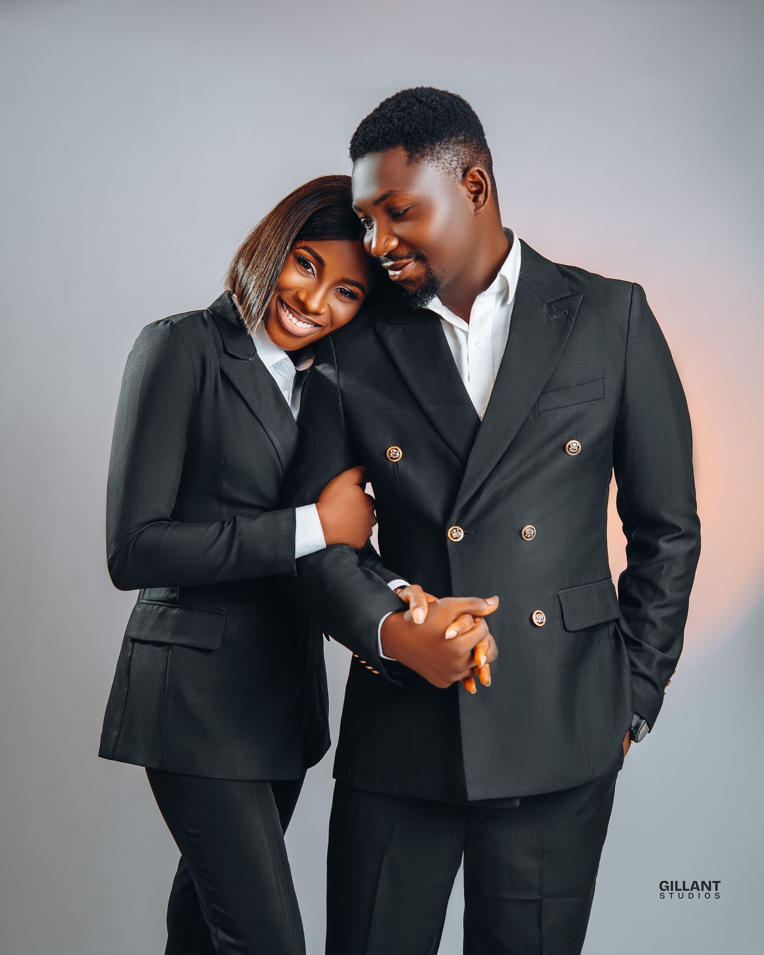Stylish couple in matching dark blazers pose intimately against studio backdrop.