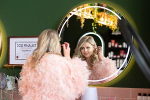 A reflection in a round mirror shows someone in a pink textured garment seated at a vanity table with lights.