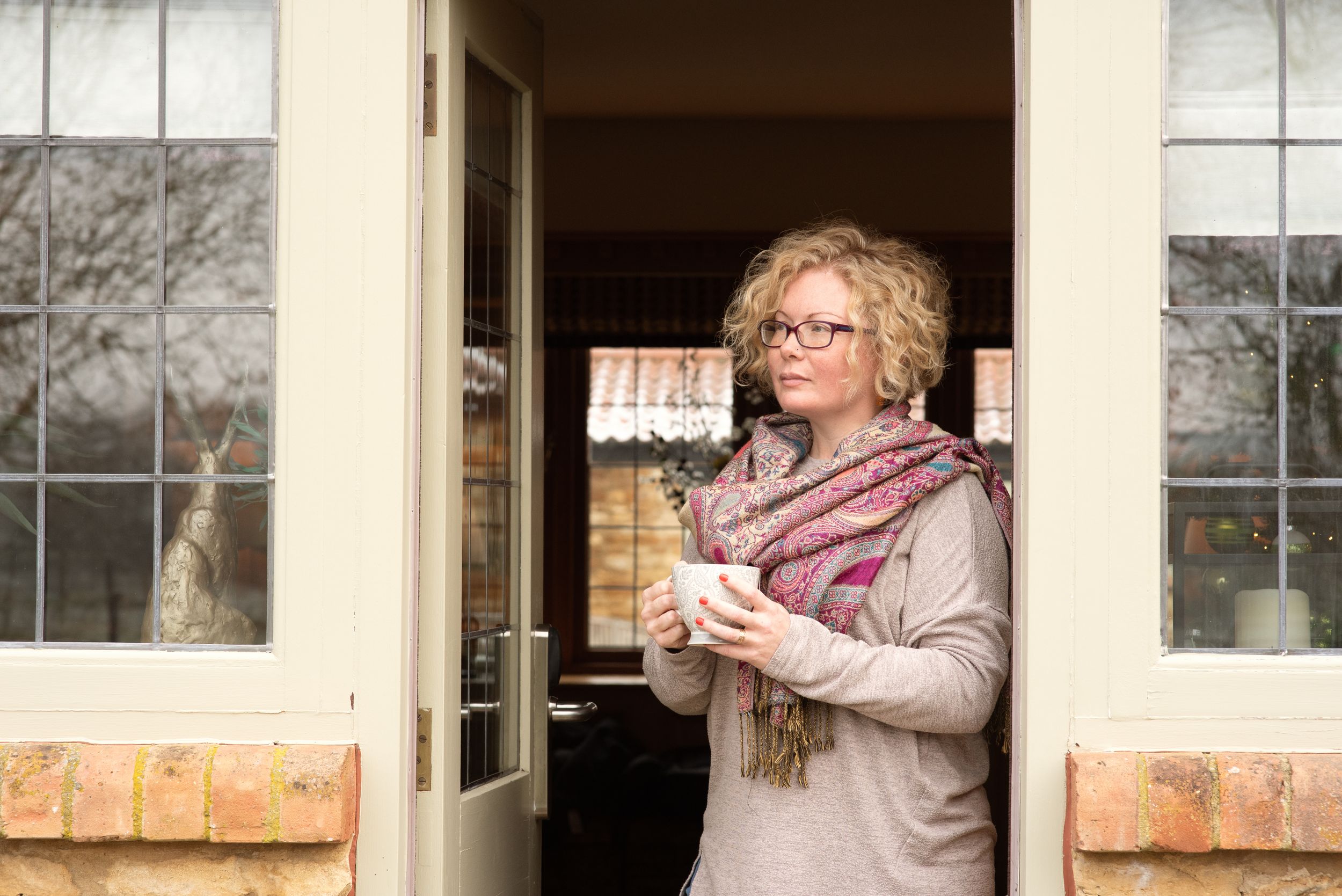 A person in glasses and a cozy scarf stands thoughtfully in a doorway between windowpanes on a winter day.