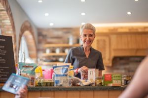A person behind a store counter smiles while surrounded by colorful retail products and snacks in a rustic shop interior.