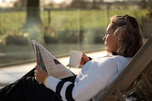 Someone relaxes in a lounge chair while reading a newspaper and holding a coffee cup in warm sunlight.