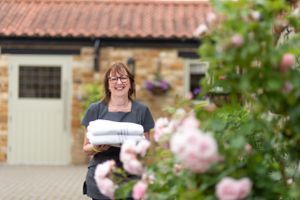 A person in a navy sweater holding white linens stands outside a stone cottage with pink roses in the foreground.