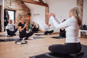 People practicing pilates stretches on exercise mats in a wooden-floored studio with exposed brick walls.