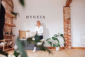 Bright minimalist wellness space with exposed brick, wooden shelving, potted plants and HYGEIA text on white wall.