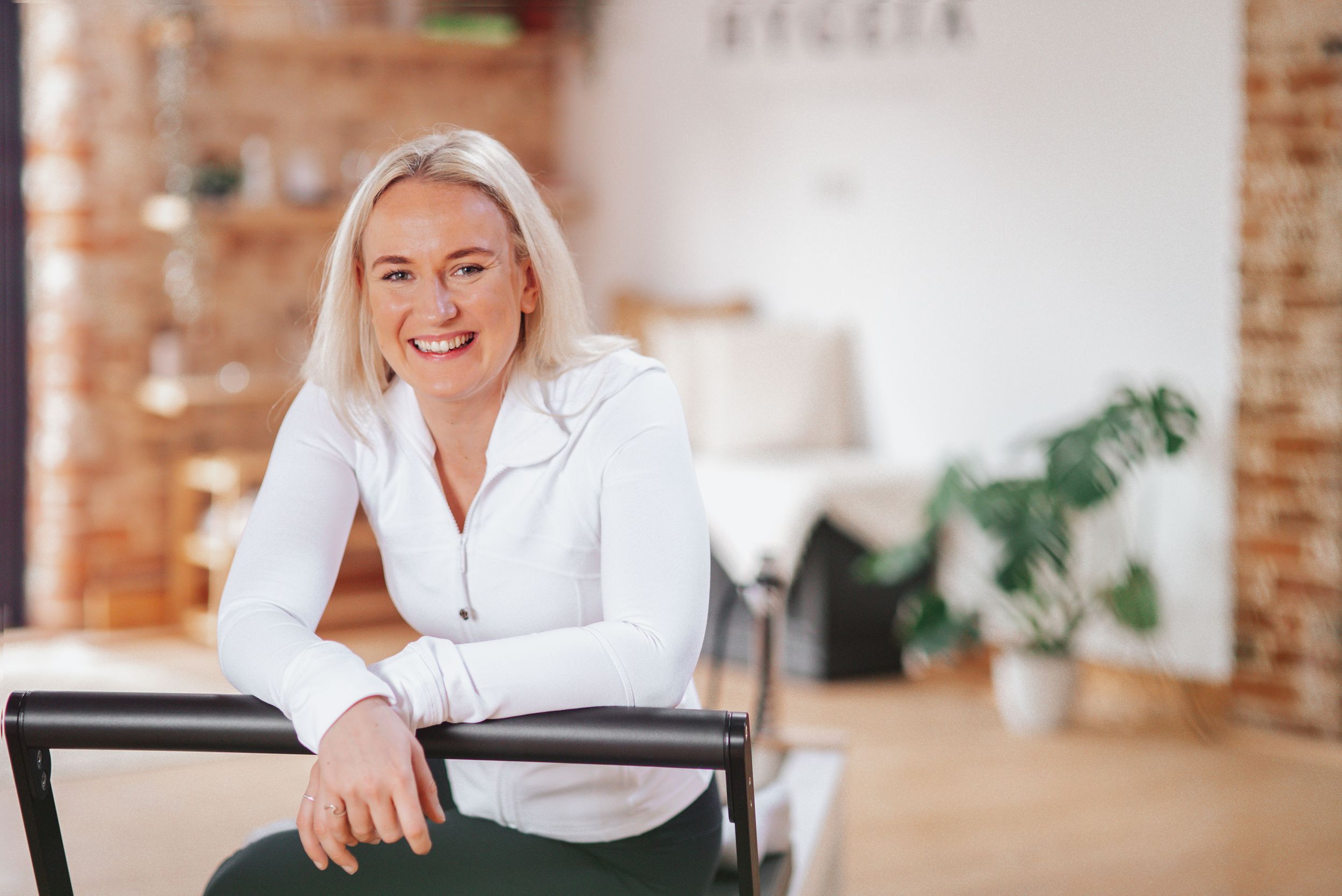 Professional in a white button-down shirt sits casually in a modern office space with exposed brick walls and plants.