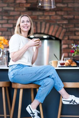 Person relaxing at kitchen counter with wine glass, wearing casual white top and jeans against brick wall backdrop.