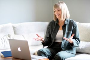 Person sitting on couch having animated conversation while working on MacBook laptop.