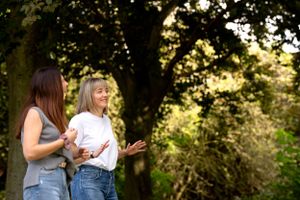 Two women in casual attire share a candid moment laughing together in a sunlit outdoor garden setting.
