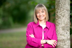A person wearing a bright pink jacket stands with crossed arms beside a tree trunk, with a blurred green background.