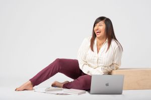 Someone sits casually on the floor in burgundy pants and white shirt, laughing while working on a MacBook laptop.