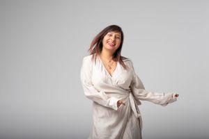 A person in a white wrap dress poses playfully against a gray studio backdrop.