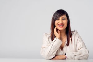 Professional wearing a beige blazer sits at white desk with a friendly expression.