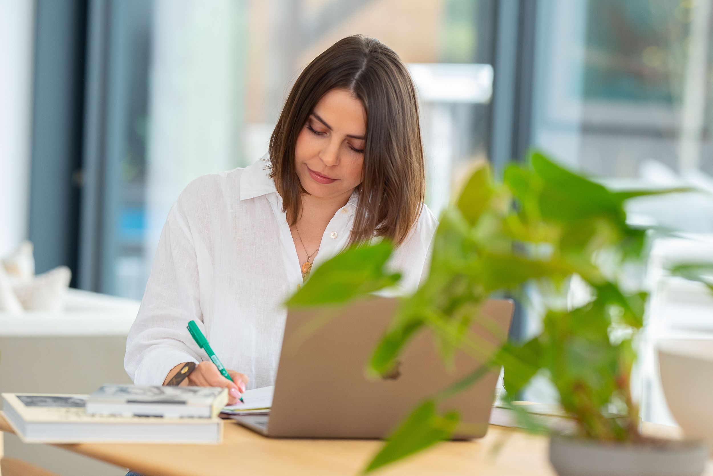 Professional in a white shirt works at a bright desk with a laptop and green plant in a modern office setting.