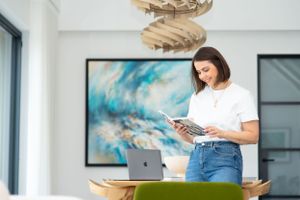 A person in casual attire stands by a laptop while reading from a digital tablet in a modern office space.