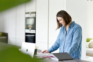 Woman working from home at kitchen counter browsing documents while using laptop in modern interior.