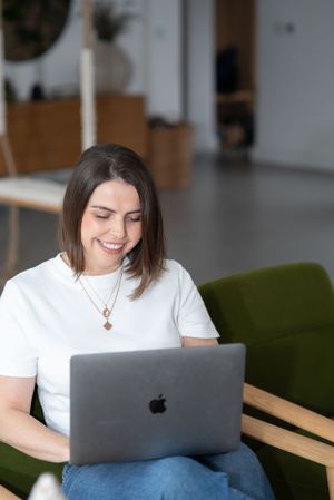 Person in white t-shirt smiling while working on MacBook laptop from green armchair.