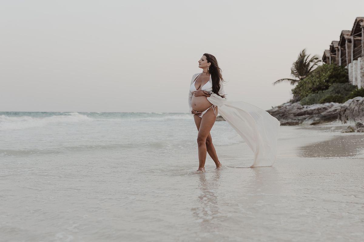 Pregnant woman in white bikini and flowing fabric walks along a tropical beach shoreline at dusk.