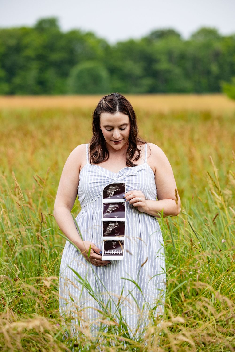 Pregnant woman in blue dress holding ultrasound photos in a golden field with green trees in the background.
