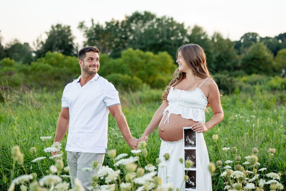 Pregnant couple holding hands in a flower field, woman in white dress showing ultrasound photo outdoors.