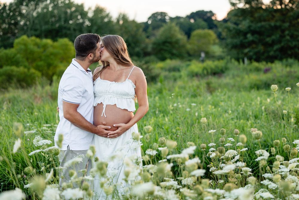 Pregnant couple kissing in a wildflower meadow, woman in white dress cradling baby bump.