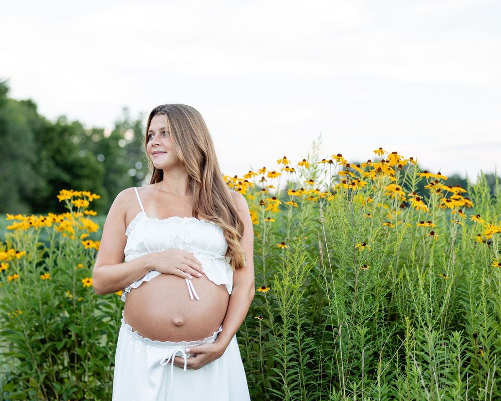 Pregnant woman in white dress standing among yellow flowers, gently holding her baby bump.