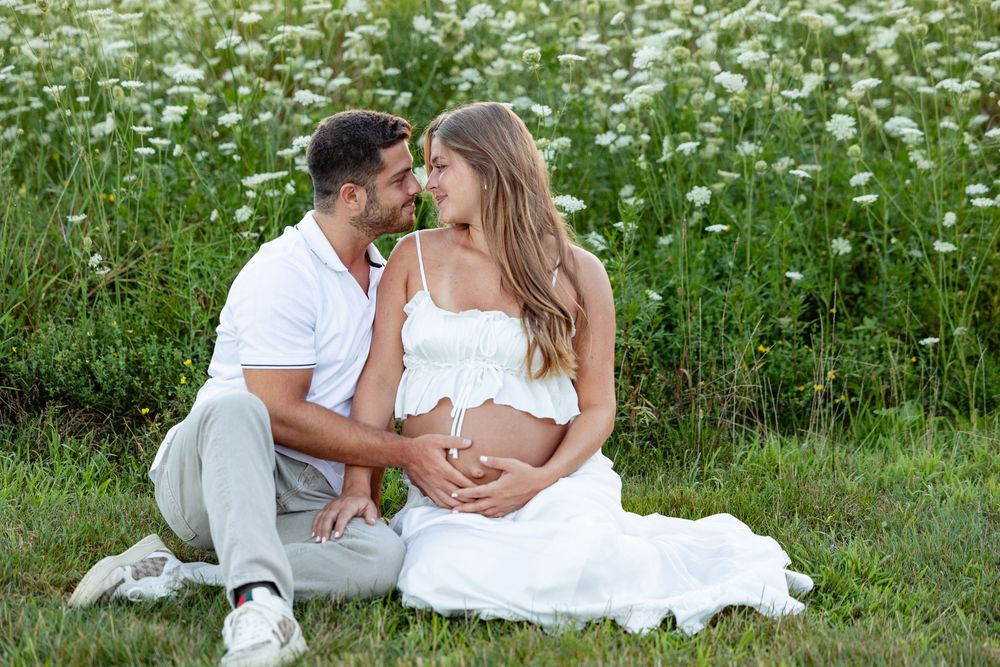 Couple sitting in a flower field, man embracing pregnant woman in white dress, sharing a tender moment outdoors.